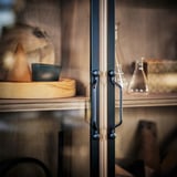 Armoire en bois avec poignées sombres, porte vitrée, exposant des objets comme une planche de bois, de la poterie et des carafes.