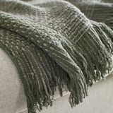 Couverture douce tissée à la main avec des franges, motif texturé gris-vert foncé, exposée sur un coussin.