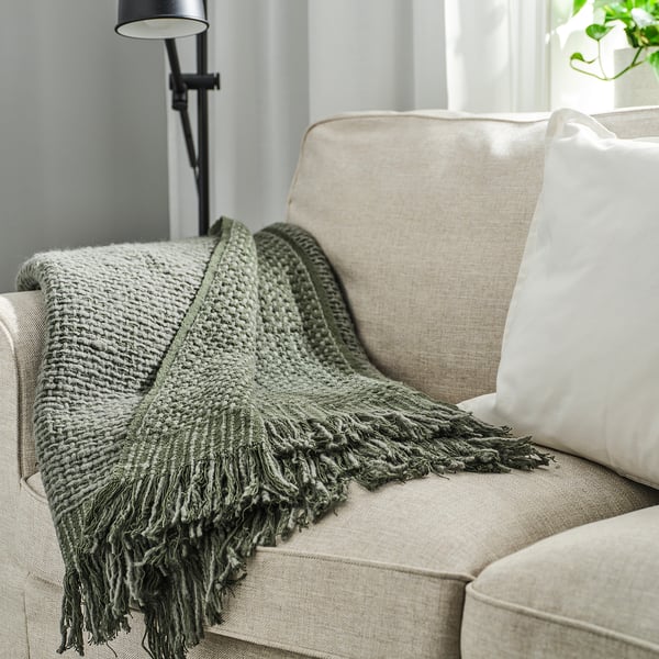 Plaid douillet LAPPTÅG en tricot gris-vert foncé sur canapé beige. Extrémités frangées, aspect texturé. Grande assise et partie du dossier.