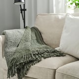 Plaid douillet LAPPTÅG en tricot gris-vert foncé sur canapé beige. Extrémités frangées, aspect texturé. Grande assise et partie du dossier.