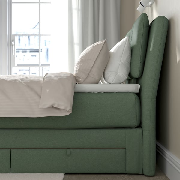 Lit avec tête de lit rembourrée verte, oreillers beiges, draps blancs et tiroirs sous le lit.