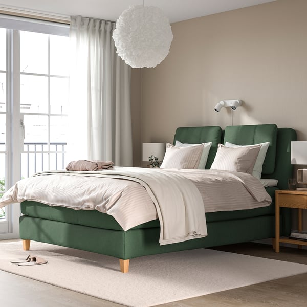 Chambre moderne avec lit LILLFJÄLLET vert et draps blancs, table de chevet en bois, tapis discret, grande fenêtre et suspension blanche.