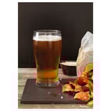 Le verre transparent LODRÄT, au design incurvé, retient la bière mousseuse, avec des éclats sur ardoise noire.