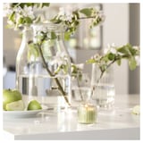 Bougie en verre transparent, cire verte, mèche allumée, pommes, fleurs dans des vases. Cadre propre et frais.