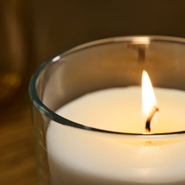 Gros plan d’une bougie blanche parfumée allumée dans un récipient en verre transparent, montrant un matériau de qualité et une flamme douce.