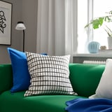 Salon lumineux avec canapé vert, oreillers bleus, coussin à carreaux, lampe blanche et plantes. L’oreiller MÄSTERROT en grille noir/blanc est proéminent.