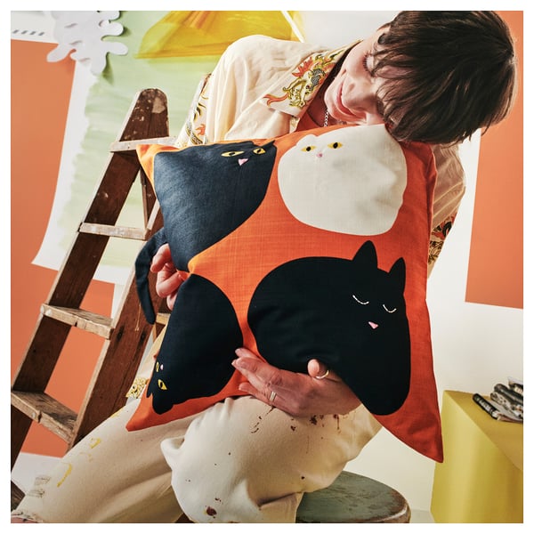 Une personne tient un coussin rouge avec des chats brodés en noir et blanc, vêtue d’une chemise multicolore.