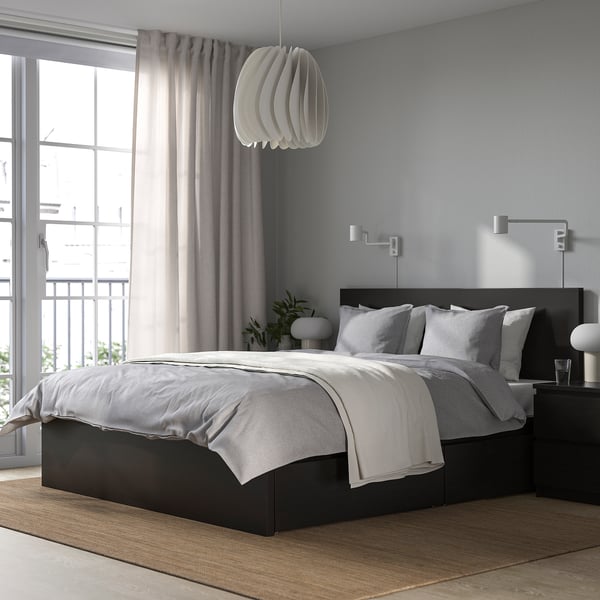 Chambre moderne avec lit noir MALM, draps gris, lampe suspendue, grande fenêtre, rideaux gris, table de chevet et plantes. Le lit est central et grand.