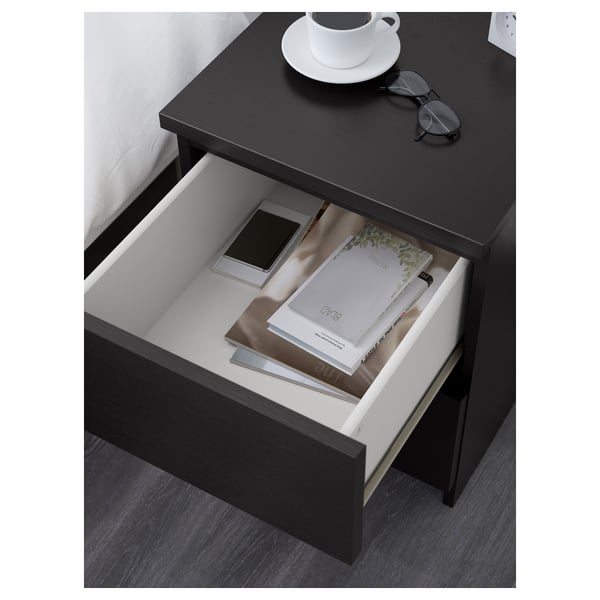 Table de chevet noire avec un tiroir ouvert sur lequel sont affichés un magazine et un téléphone, placée à côté d’un lit.