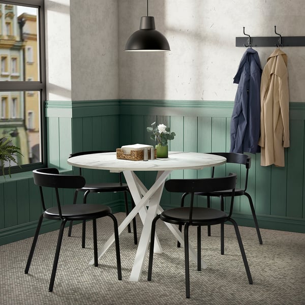 Chambre confortable avec table mariam et chaises noires près de la fenêtre. Deux manteaux pendent, le luminaire ajoute une touche moderne.