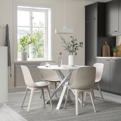 Table à manger ronde moderne avec cadre en acier blanc, pieds entrecroisés, chaises assorties et tapis rayé près d’une grande fenêtre.