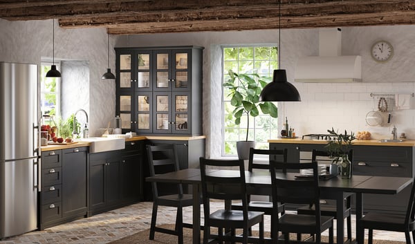 Cuisine rustique moderne : armoires noires, comptoir en bois, réfrigérateur en acier inoxydable, grande fenêtre, lampes suspendues, chaises en bois, dosseret carrelé, horloge.