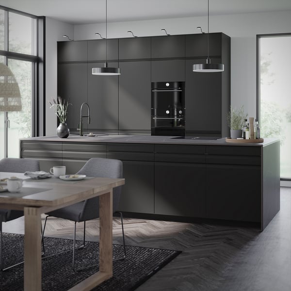 Cuisine moderne avec armoires mates gris foncé, four encastré et table à manger en bois avec chaises. De grandes fenêtres laissent entrer la lumière naturelle.