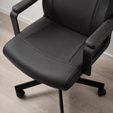 Chaise de bureau noire avec assise et dossier rembourrés, hauteur réglable, accoudoirs et roulettes, positionnée sur un plancher en bois.