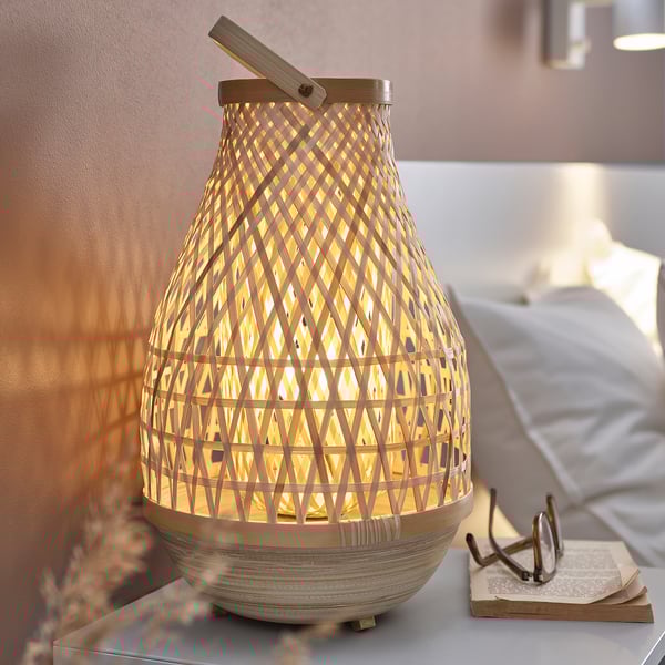 Une lampe MISTERHULT avec un cadre en bambou beige naturel, dotée d’une lumière chaude et rougeoyante, posée sur une table de chevet.
