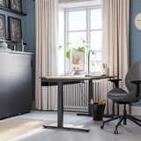Bureau à domicile moderne : le bureau noir MITTZON s’adapte parfaitement sous une grande fenêtre, une chaise grise, des rideaux beiges, des plantes, des étagères et un mur bleu.
