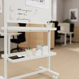 Unité mobile blanche avec des étagères, des marqueurs et une tablette. Comprend un tableau blanc avec des notes manuscrites, dans un bureau.