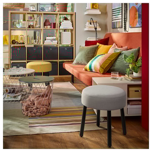 Salon coloré : pouf gris, canapé orange, tabouret jaune, tapis multicolore, étagères en bois avec décor.