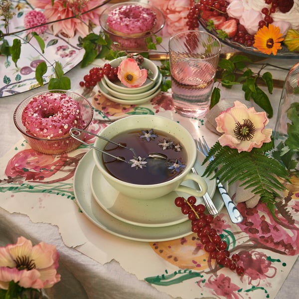 Table dressée avec de la vaisselle TRÈS MALSAINE : tasse de thé, beignets, eau, fleurs et couverts.