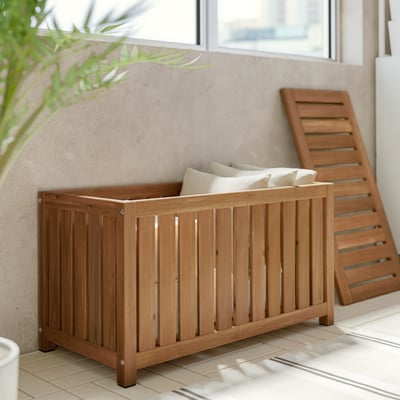 Une boîte de rangement extérieure NÄMMARÖ en bois d’acacia brun avec un design à lattes pour la ventilation et une double fonctionnalité en tant qu’assise.