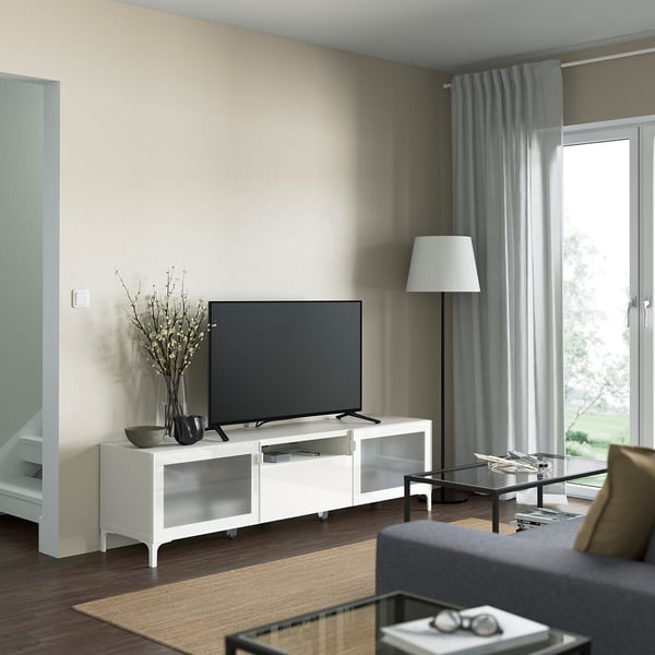 Salon avec meuble TV NANNARP blanc, télévision noire, lampadaire, rideaux gris, canapé gris et table basse en verre.