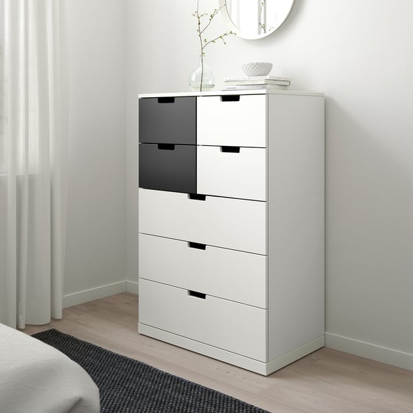 Une commode NORDLI blanche. Il est grand et étroit, s’insérant parfaitement dans un coin. Le dessus a des objets décoratifs et les tiroirs sont modernes et élégants.