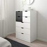 Une commode NORDLI blanche. Il est grand et étroit, s’insérant parfaitement dans un coin. Le dessus a des objets décoratifs et les tiroirs sont modernes et élégants.
