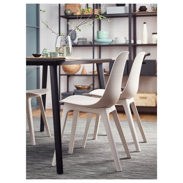 Scène de salle à manger moderne avec des chaises ODGER beige clair autour d’une table sombre. Des étagères en arrière-plan contiennent divers articles.