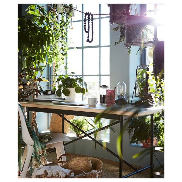 Chambre lumineuse et ensoleillée avec un bureau en bois près d’une grande fenêtre. Le bureau contient des plantes, une cafetière et des livres. Panier tressé et chaise blanche à proximité.