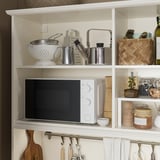 Kitchenette blanche avec micro-ondes, râpe, bols, bocaux et serviettes ; Stockage ouvert et fermé.