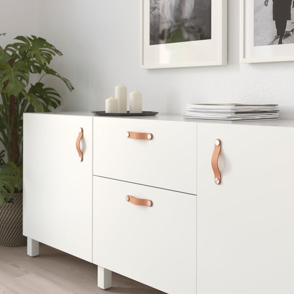 Armoire blanche avec poignées en cuir ; Comprend deux portes et deux tiroirs, avec des objets décoratifs sur le dessus.