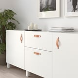 Armoire blanche avec poignées en cuir ; Comprend deux portes et deux tiroirs, avec des objets décoratifs sur le dessus.