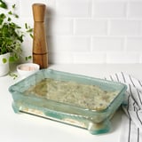 Plat de cuisson en verre avec une teinte verte, contenant des aliments cuits, placé sur un tissu rayé, à côté de la plante, d’un bol de sel et d’un moulin à poivre.