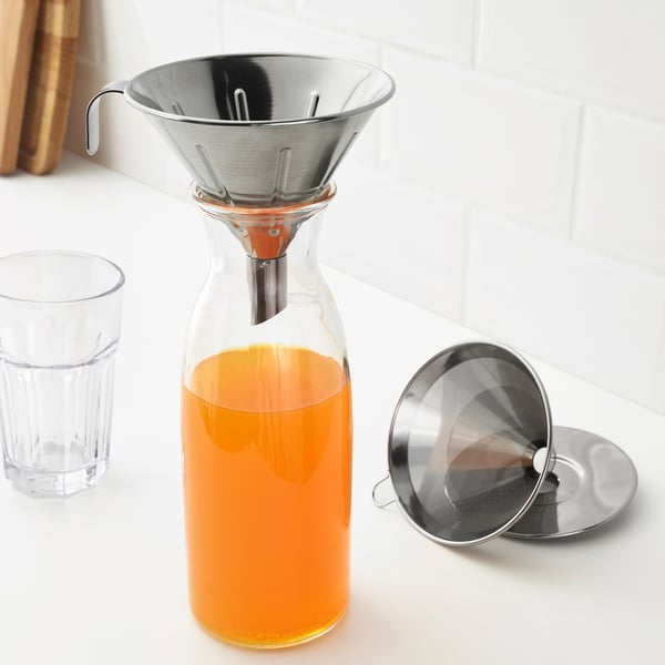 Carafe en verre avec liquide orange, filtre métallique sur le dessus, pièce filtrante supplémentaire à côté.