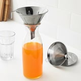 Carafe en verre avec liquide orange, filtre métallique sur le dessus, pièce filtrante supplémentaire à côté.