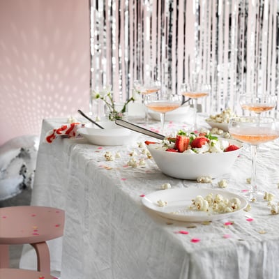 Dressage de table avec bols et verres OFTAST, verre trempé blanc, maïs soufflé éparpillé, fraises et confettis roses, fond argenté brillant.