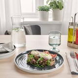 Une table rustique avec une assiette en céramique contenant une salade et des tomates tranchées. Des verres avec de l’eau, une carafe et des bouteilles d’huile sont posés sur la table.