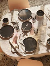 Dressage de table OMBONAD : assiettes en céramique noire et blanche, bols, verres, serviettes en bois, nappe naturelle.