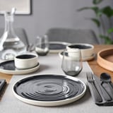 Configuration moderne de la table OMBONAD ; Assiettes en céramique noir et blanc, tasses en verre, bol en bois, couverts gris.