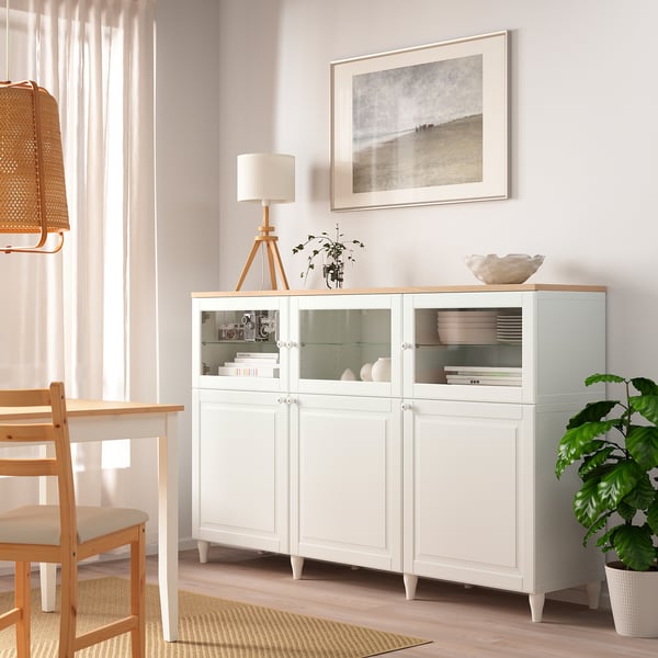 Armoire OSTVIK blanche avec portes vitrées dans un salon minimaliste, flanquée d’une lampe et d’une plante.