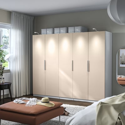 Chambre moderne avec grande armoire beige, charnières à fermeture douce, éclairage intérieur, banc marron avec des vêtements, miroir rond.