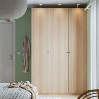 Chambre moderne avec armoire PAX en bois clair, mur vert, porte blanche, éclairage intégré et lit d’édredon rayé.