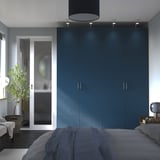 Armoire PAX avec 4 portes à côté de la porte vitrée de la salle de bain. Poignées en argent, lit avec couette grise, plante d’intérieur et lampe.