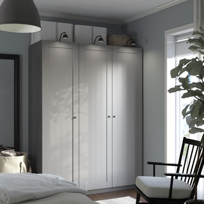 L’armoire PAX grise moderne se dresse fièrement à côté de la fenêtre, flanquée d’une chaise et d’un lit, avec un design élégant et de multiples compartiments.