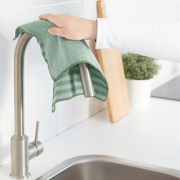 Personne tenant un chiffon de nettoyage vert sous un robinet de cuisine, avec une planche à découper et une plante en arrière-plan.