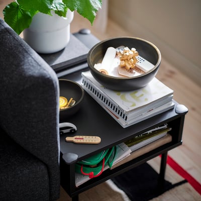 Petit bol en bois d’acacia sur une table d’appoint, contenant des pièces de jeu, à côté d’un livre et d’une petite plante, près d’un fauteuil gris foncé.