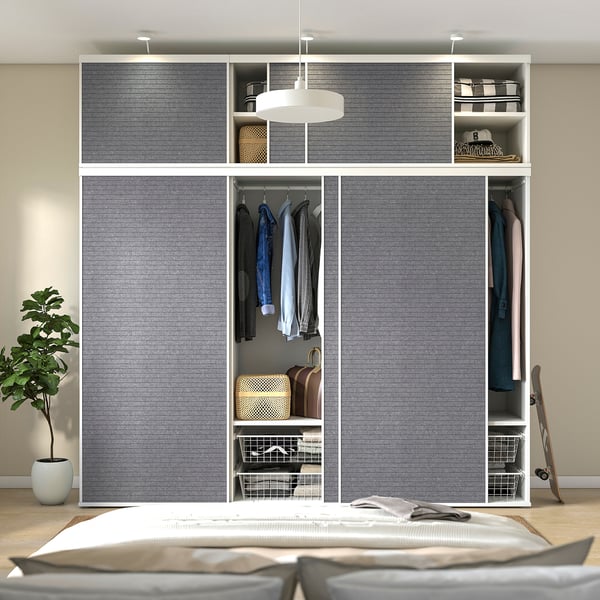 Placard de chambre avec portes coulissantes, vêtements suspendus, étagères et luminaire. Plante à gauche, lit au premier plan.