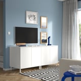 Salon moderne avec murs bleus, rangement PLATSA, télévision, tapis rayé, rideaux gris et cadres d’art.