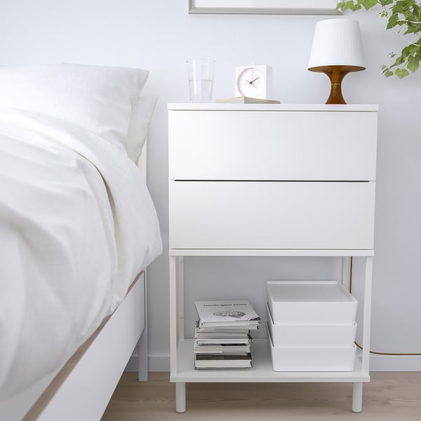 Une table de chevet blanche PLATSA au design moderne, dotée d’un tiroir, d’une étagère ouverte et d’objets de décoration blancs.