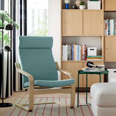 Fauteuil POÄNG IKEA bleu près d’une étagère, sur tapis. Table verte avec des magazines et un casque, un lampadaire à proximité. Espace de lecture douillet.